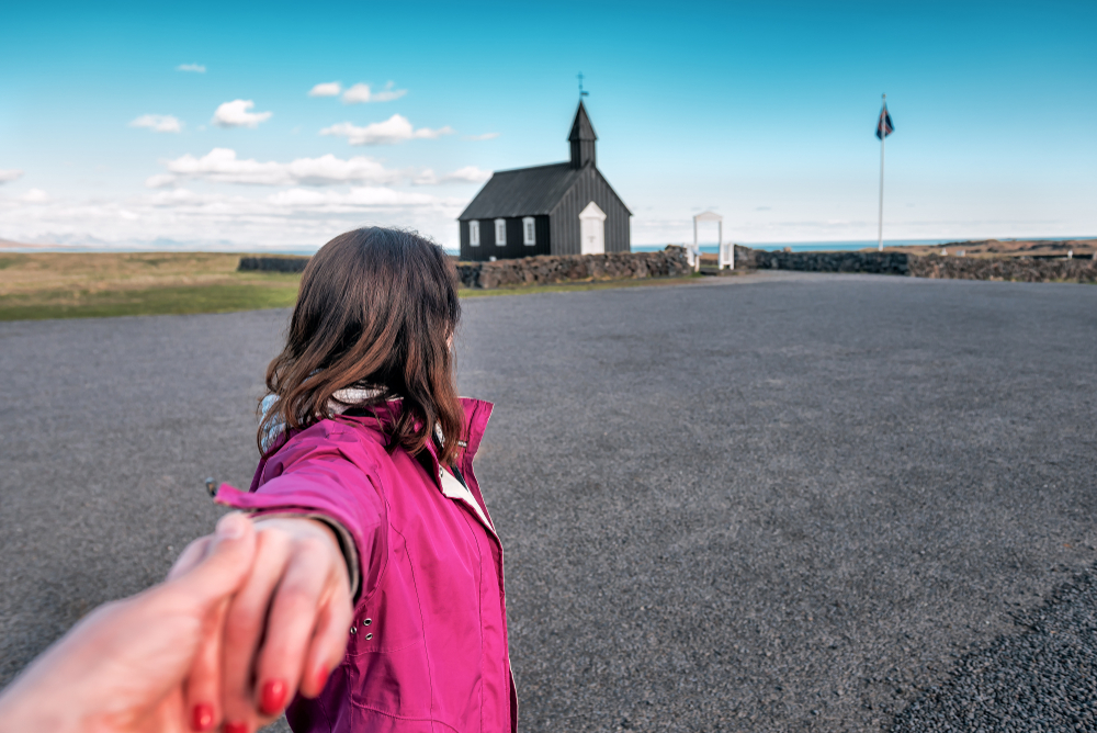 A woman in a pink windbreaker holds the hand of a man off camera as she faces the black church in Iceland.