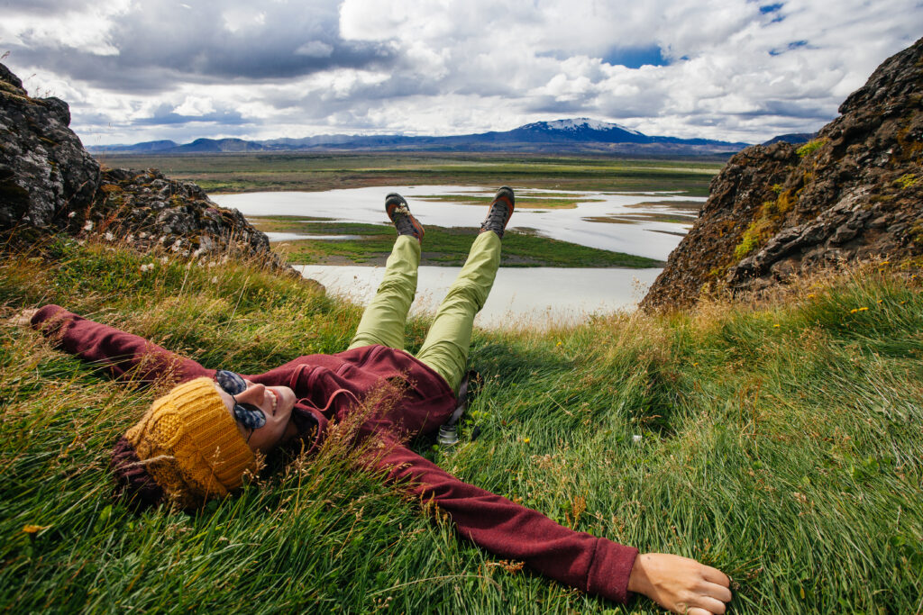 A woman lounges in the green grass of Iceland in a windbreaker, hiking pants, beanie and sunglasses.