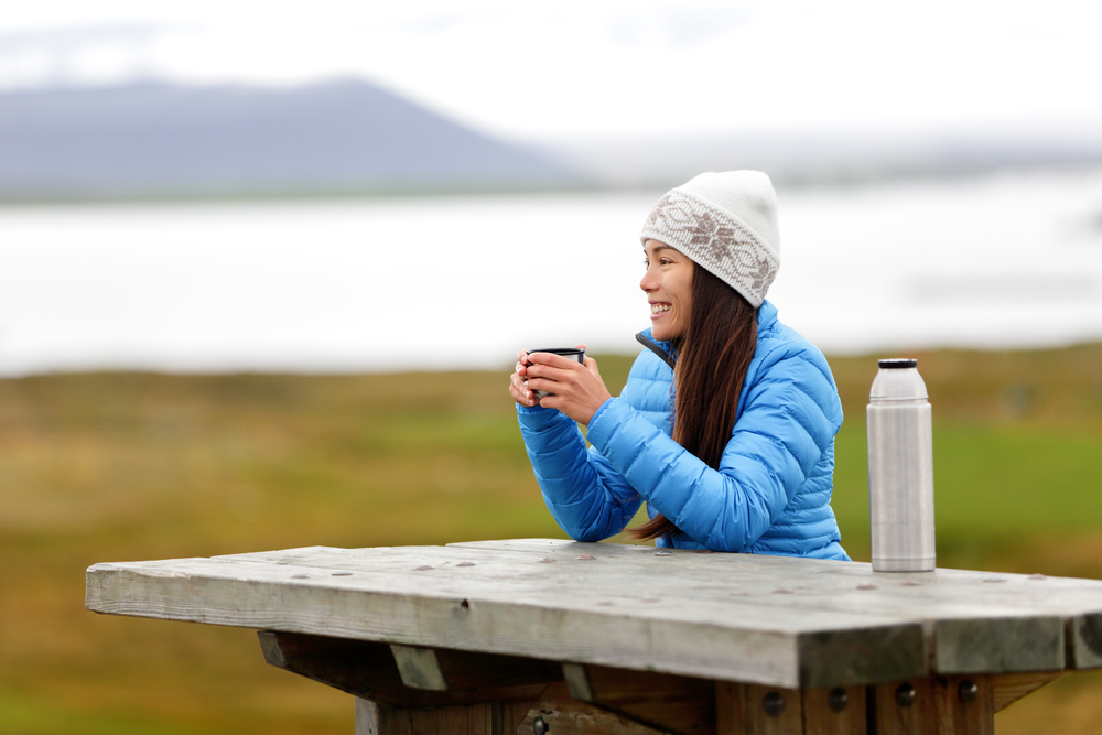 A woman takes a break at a picnic table and enjoys the views in her beanie and blue puffer jacket as she sips hot coco from a thermos.