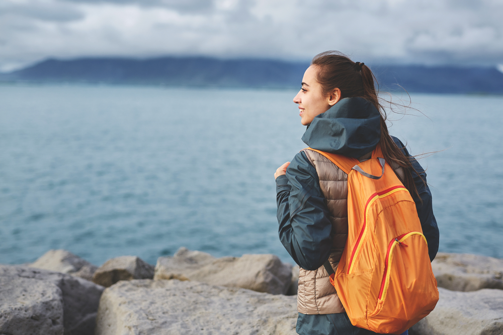 A woman looks over the cool cliffs, not worrying about what to wear to Iceland in July, as she is comfortable and cool in her blue windbreaker, orange pack, and puffer jacket.