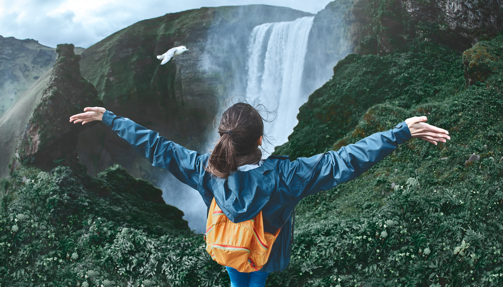 A woman knows what to wear to Iceland in July when she's on a hike: her orange backpack and blue windbreaker stand out against the green foliage and the water of the waterfall!