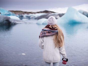 woman standing in front of lake with giant pieces of icebergs in the background. she is wearing a white beanie and has blonde hair. she is wearing a white long sweater with a colorful scarf and holding a red coffee mug to her right side. she is facing the lake