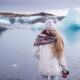 woman standing in front of lake with giant pieces of icebergs in the background. she is wearing a white beanie and has blonde hair. she is wearing a white long sweater with a colorful scarf and holding a red coffee mug to her right side. she is facing the lake