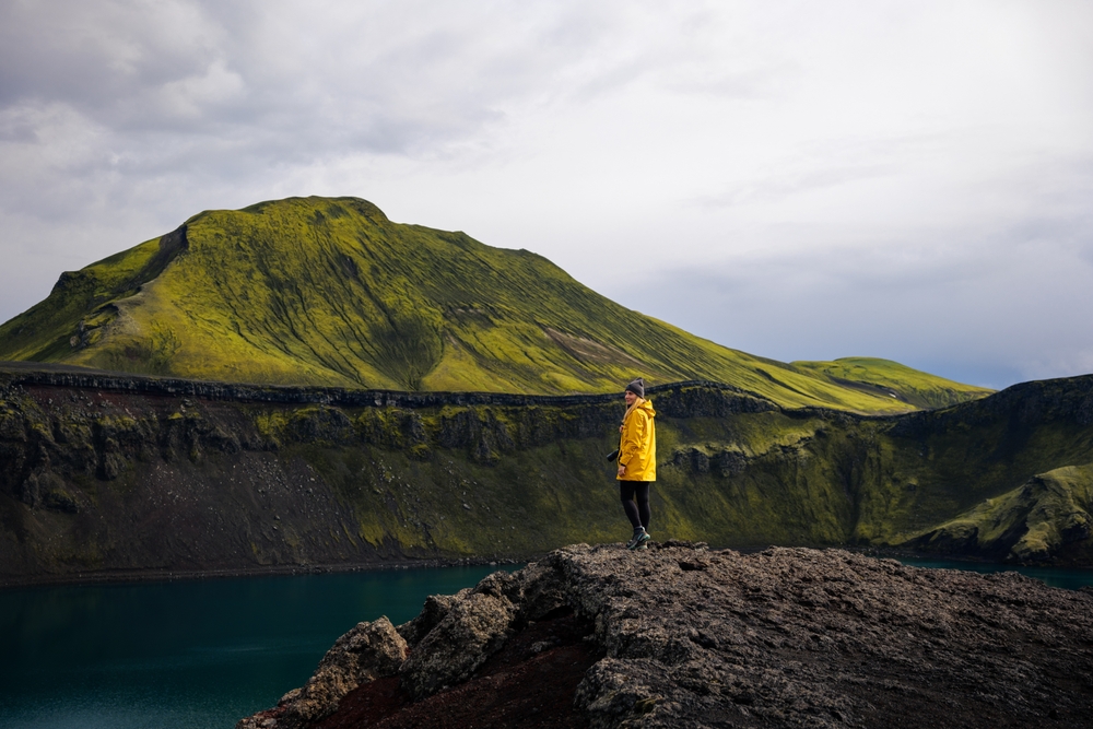 A woman in black leggings, boots, a beanie and a yellow rain jacket knows what to wear in Iceland in June: she is ready to layer up as needed, which is now as she navigates toward the water on the edge of a rock! 