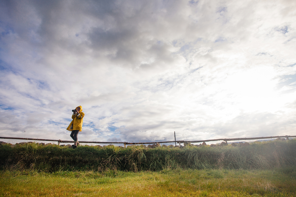 A woman in a yellow rain coat points her camera off screen as she stands on the greens of the Highlands: her hiking boots and fleece lined leggings are keeping her warm. 