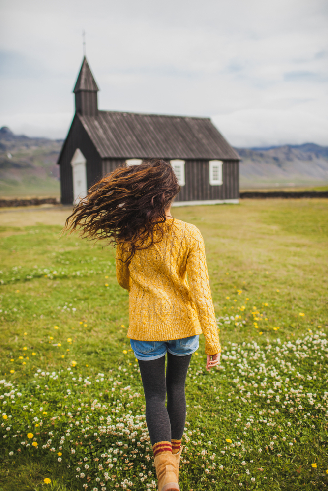 A woman walks toward a black church, her curly hair swinging behind her, as she wears a yellow knit sweater,  shorts, fleece leggings, and boots. 