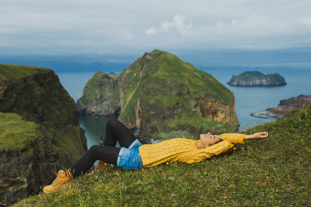 A young woman knows exactly what to wear in Iceland in June: her yellow knit sweater keeps her warm as she lays down in the grass, jean shorts, leggings and boots comfortable and stylish. 