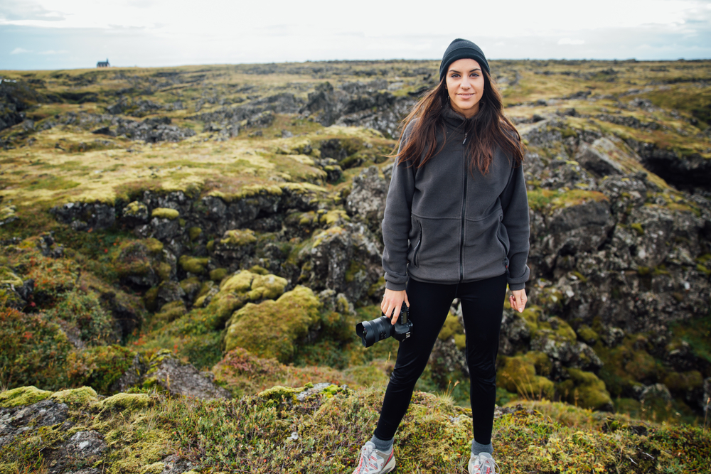 A young woman with dark brown hair wears hiking pants, sneakers, a fleece jacket and a beanie: she holds a camera, ready to go. 