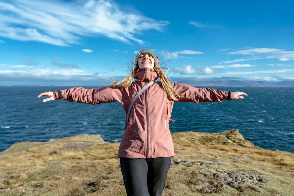 This young woman knows what to wear in Iceland in June: her hiking pants, cross body bag,  and windbreaker keep her warm as she outstretches her arms and basks in the sun. 