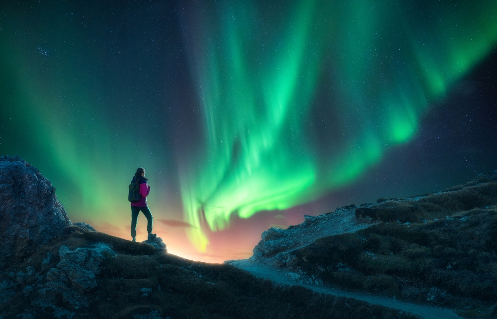 A hike stands on a mountainside looking up at the green hues of the Northern Lights.