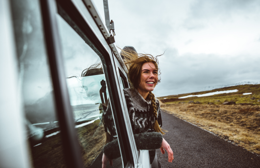 A woman with a braid and Nordic sweater hangs her head out of a car to admire the landscape: she knew things people forget when packing for iceland and is prepared for it.