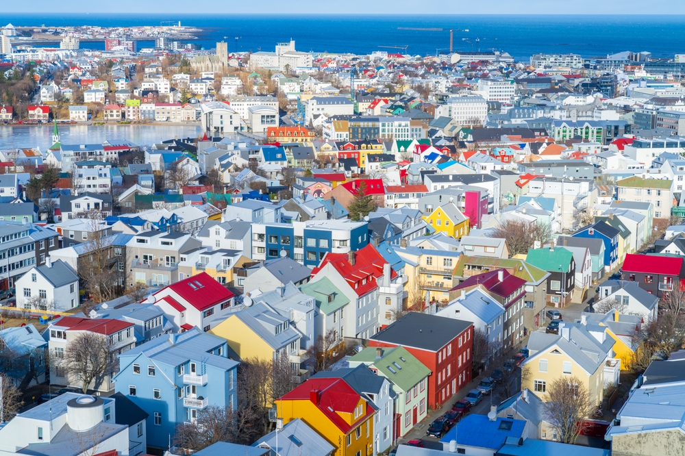 An arial shot of a town of Iceland shows brightly colored buildings all together and the cusp of the blue coast beyond.