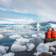 Two people in orange windbreakers sit on a rock observing the icy layers in the water on the coast: they are warm because they knew what things people forget when packing for Iceland and avoided it!