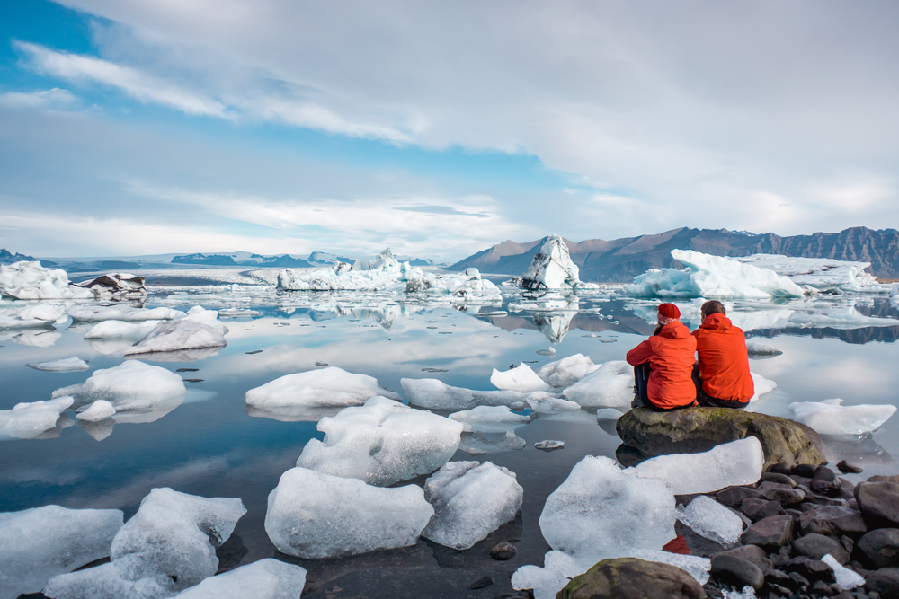 Two people in orange windbreakers sit on a rock observing the icy layers in the water on the coast.