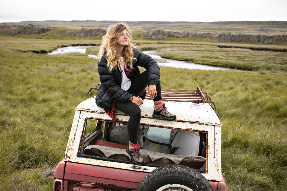 A young woman in glasses and a puffer jacket sits atop a jeep and admires the world around her.