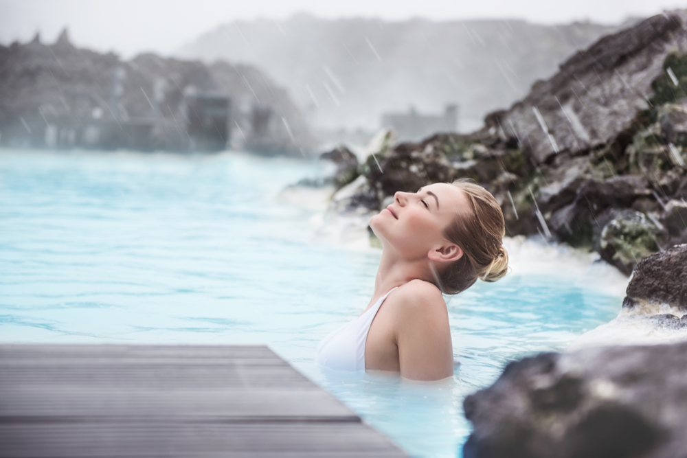 A woman leans back and enjoys the crystal blue water of a lagoon, her hair up in a bun, as she was prepared because she knew things people forget when packing for iceland