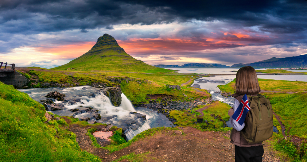 A woman with a backpack overlooks the green hills and sunset and falls in Iceland because she knew things people forget when packing for iceland and was prepared.