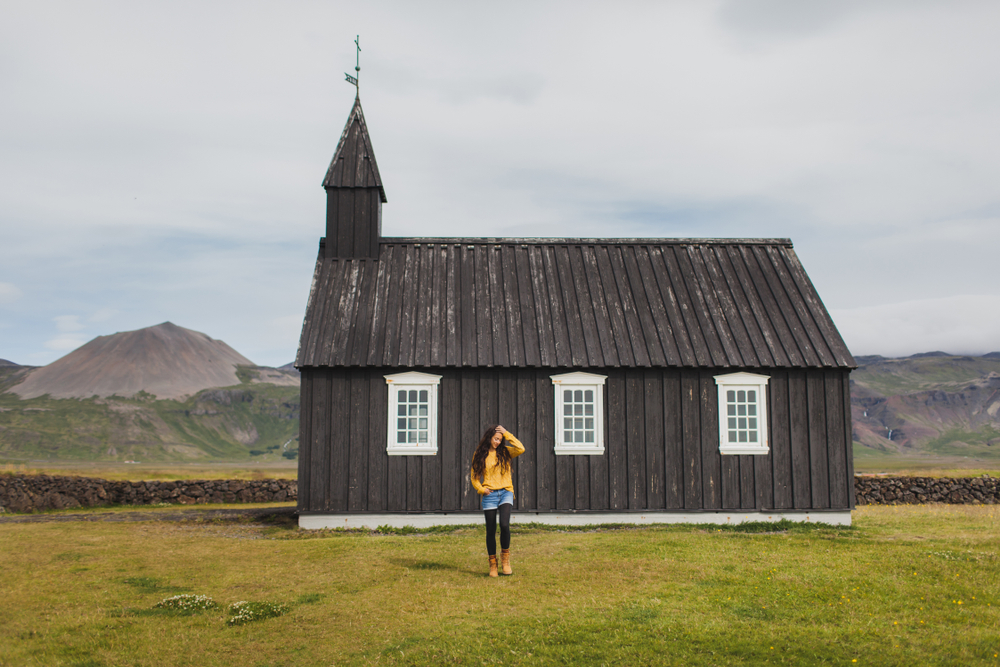 A woman in a yellow sweater, leggings, jean shorts and boots stands in front of a famous black church in the rugged landscape of Iceland.