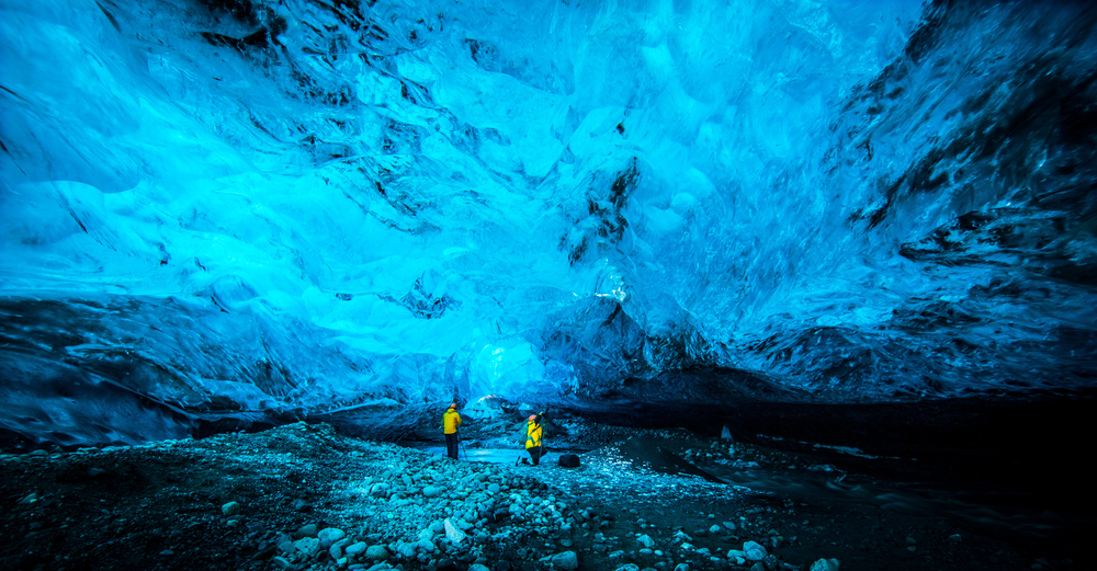 Two tourists go under the ice caves in Iceland, well prepared since they knew things people forget when packing for iceland, and explore the depths of the blue world in their yellow jackets.