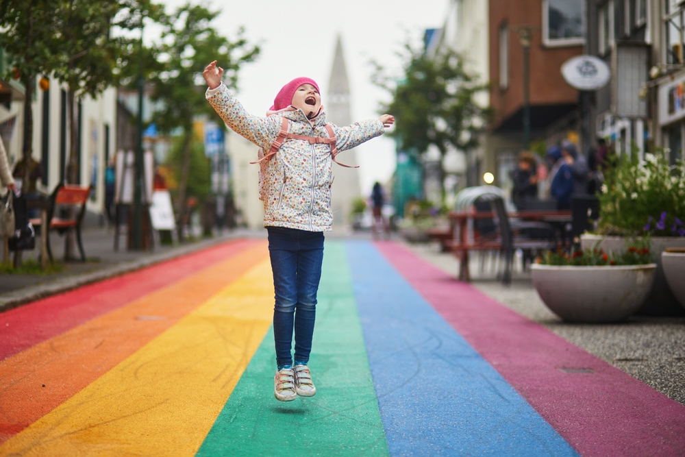 A young kid jumps up in excitement at the rainbow walkway of a city in Iceland, her jeans, pack and sweater keeping her warm.