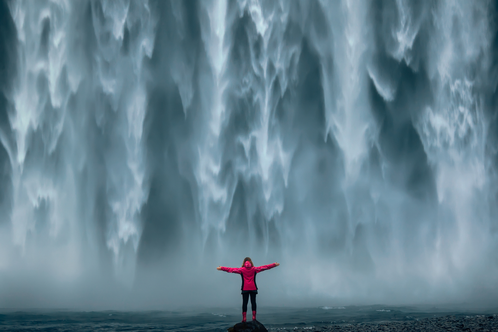A woman in a red windbreaker and boots stands with her arms spread out in front of huge falls.