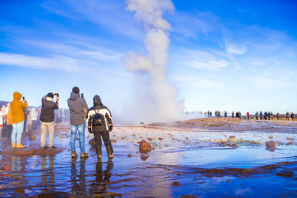 A group of tourists film a water spout on the edge of the land in Iceland, all wearing jeans or hiking pants and windbreakers and beanies: they knew what to wear to Iceland in the fall.