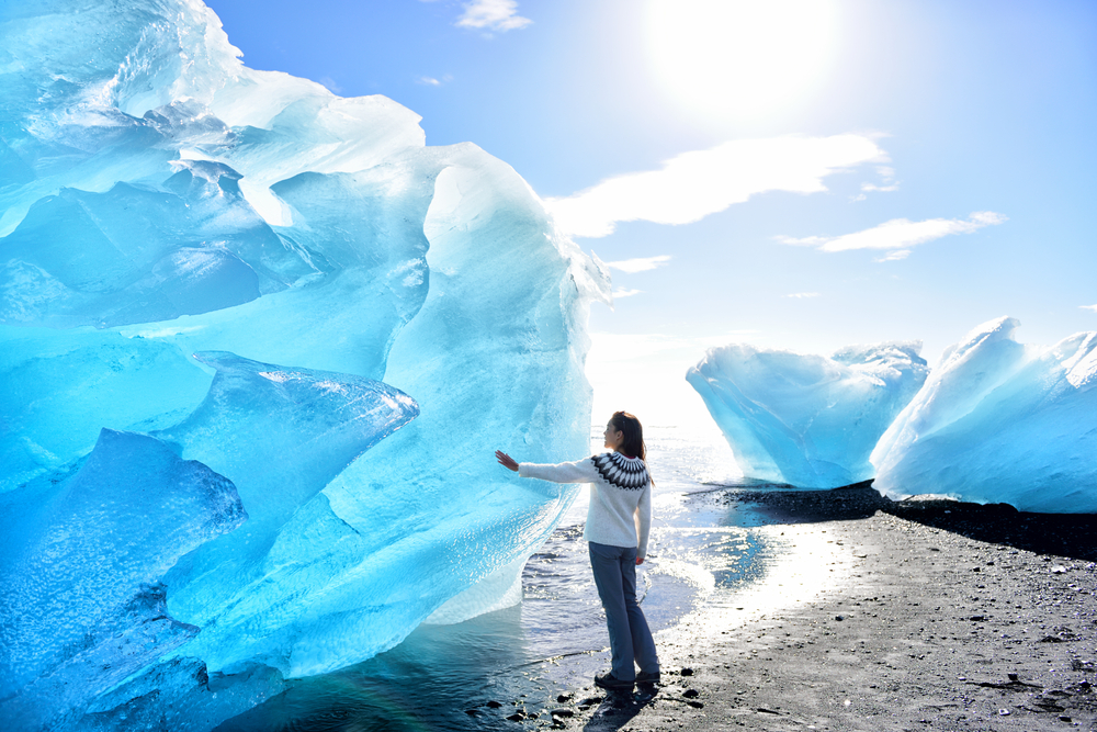 A woman stands on black sand and touches a blue glacier while wearing hiking pants, boots, and a knit sweater: she knew what to wear to Iceland in the fall.