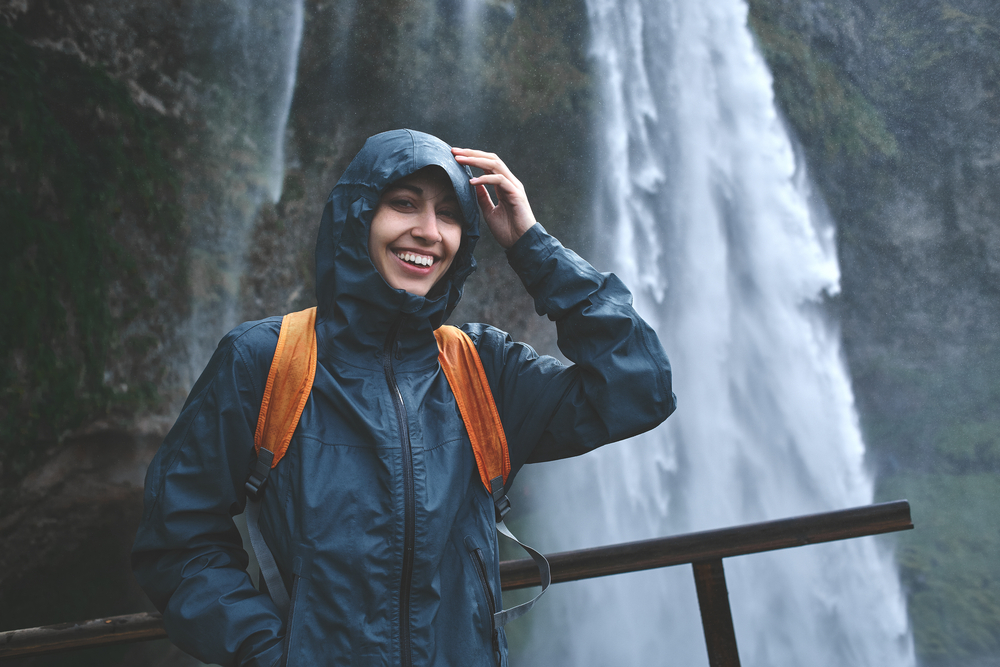 A woman in her windbreaker smiles at the camera with her hood up to protect her and her softshell orange backpack from the mist.