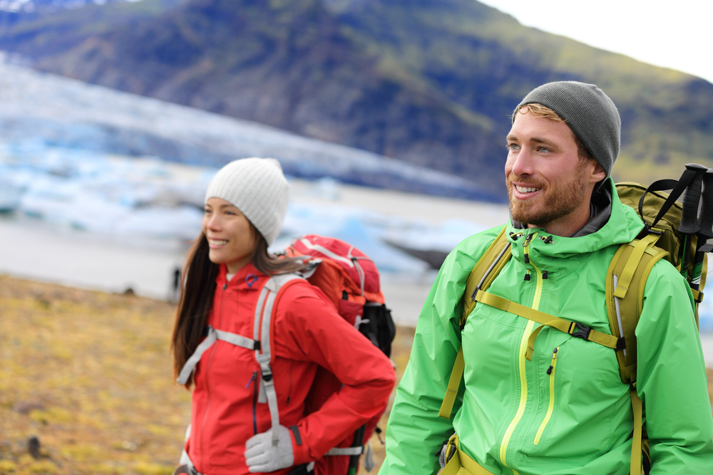 Two hikers, a girl and a guy, walk through the fall landscape in windbreakers, gloves and beanies, with softhsell backpacks on.