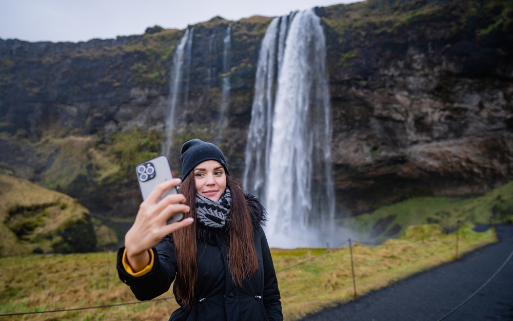 A woman takes a chilly selfie by a waterfall in a scarf, puffer jacket and beanie.