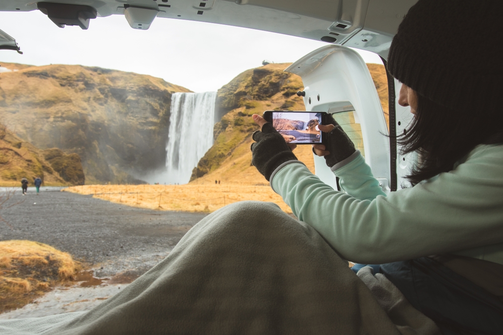 A woman sits in the back of her van and takes a photo of a waterfall while draped in a blanket with gloves on and her beanie and fleece jacket keeping her warm.