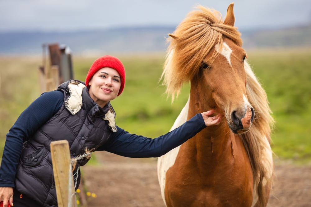 A woman pets a horse in Iceland while wearing thermal layers, a puffer jacket and a red hat, well prepared and knowledgeable about what to wear to Iceland in the fall.
