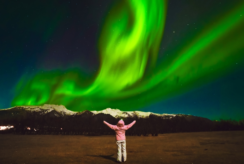 A tourist stands below the green lights of the Northern Lights, mountains ahead of them, in their hiking pants and pink parka: they knew what to wear to Iceland in the fall .