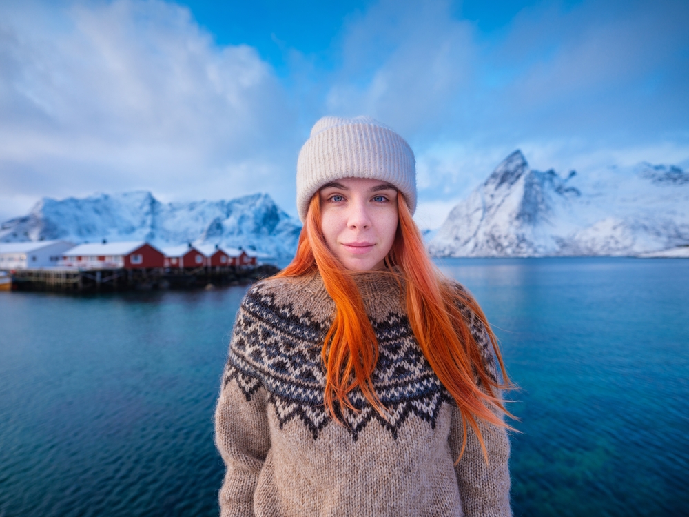 A woman with vibrant red hair and blue eyes has a beanie on and a wool sweater as she smiles at the camera, a town behind her, along with blue water and snow capped mountains.