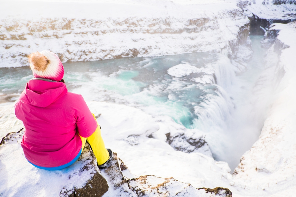 A woman in yellow hiking pants, a pink parka, and a wool beanie sits above a waterfall and looks down, comfortable since she knew what to wear in Iceland in November.