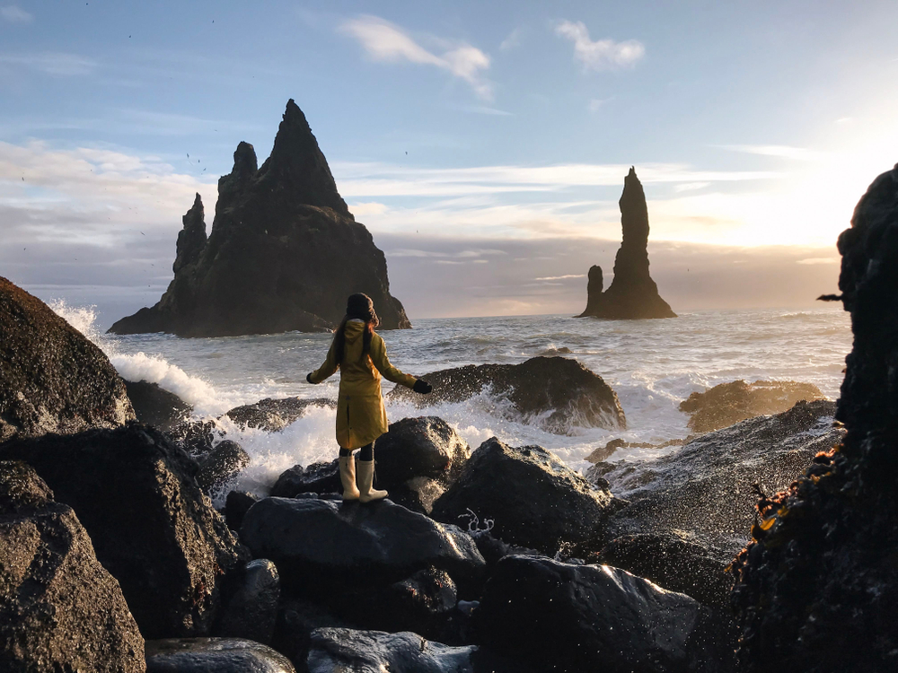 A woman in a yellow park and gloves and waterproof boots stands on the edge of a shore on a black sand beach, waves crashing over the rocks.