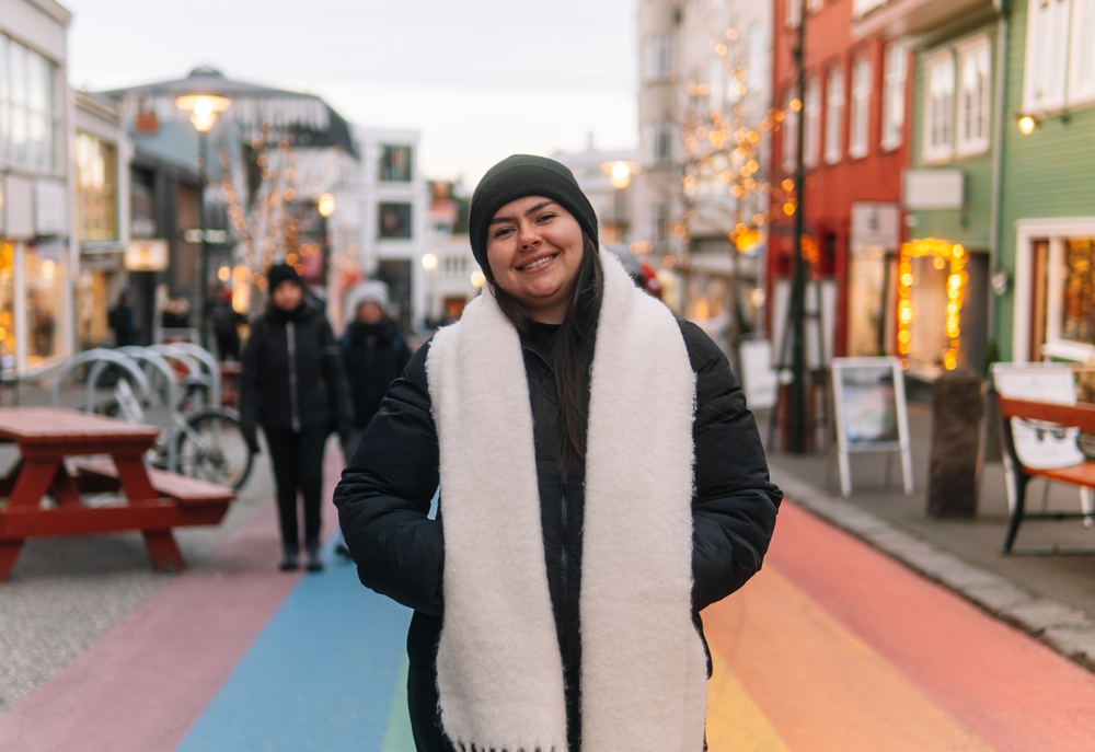 A woman smiles in the streets of Rekyjavik during the winter season in her parka, wool scarf and beanie: Christmas lights twinkle on the city's buildings behind her.