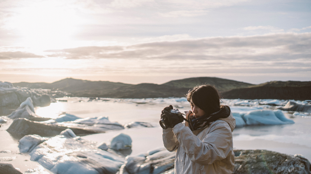A woman in gloves, and a hooded and waterproof parka raises her camera to take a picture of the icy terrain in Iceland.