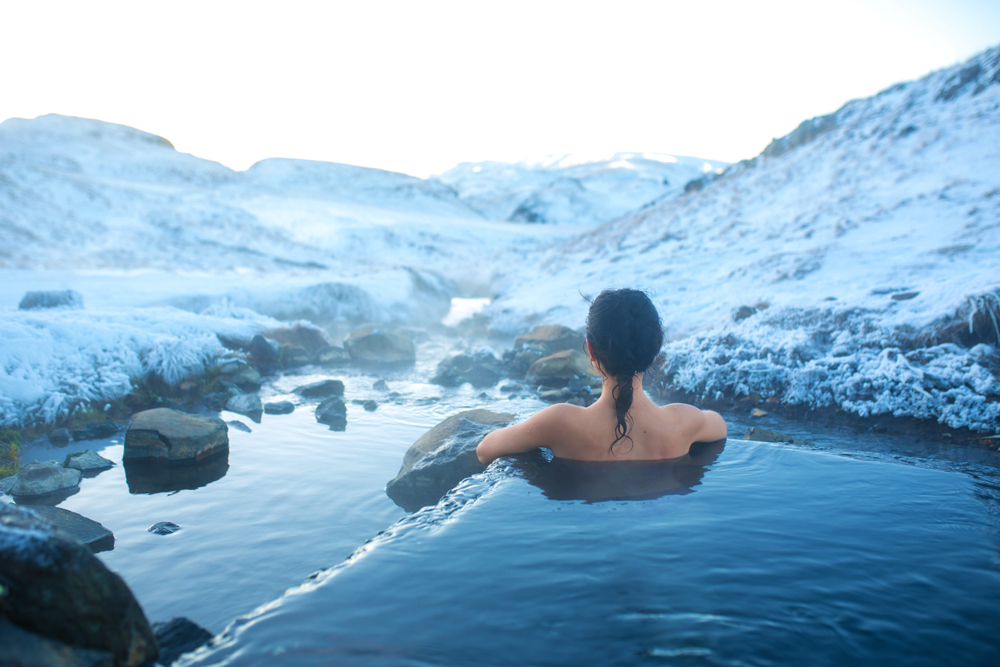 A woman sits on the edge of a hot spring in Iceland, warm in the water despite the ice mountains and rocks that surround her.