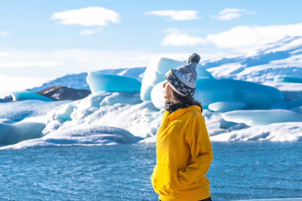 A woman in a yellow hoodie, beanie, and neck gaiter stands and breathes in the coast of Iceland with the blue waters, icy terrain and more.