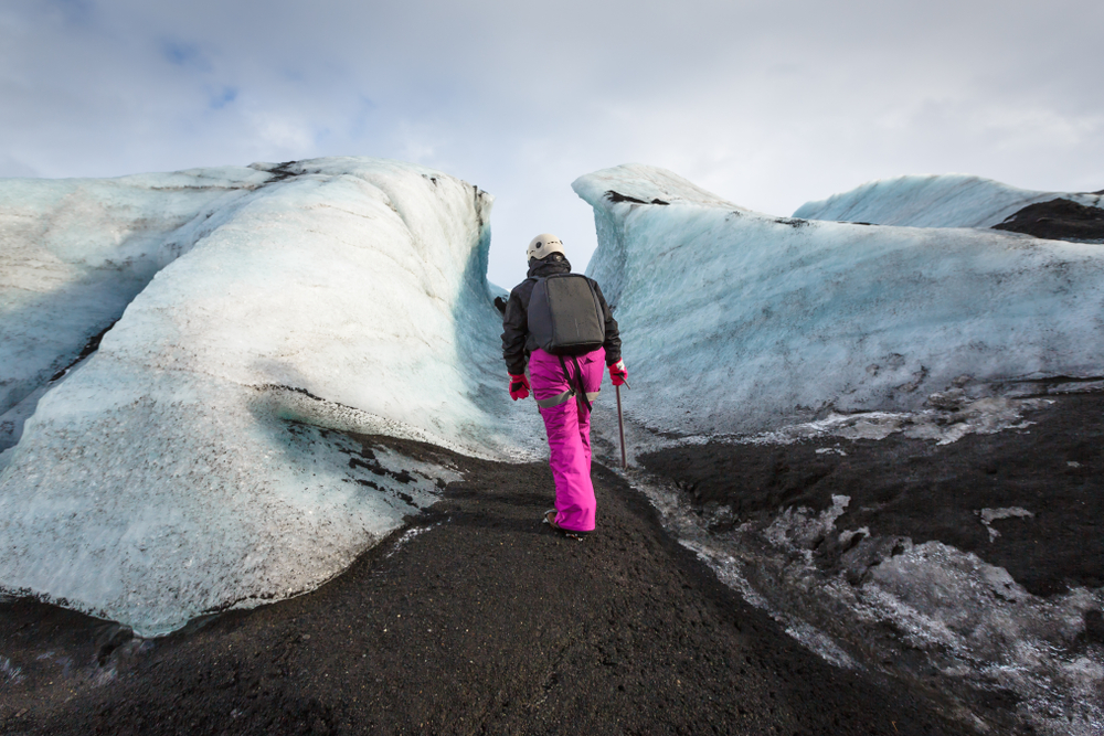 A woman in hot pink hiking pants climbs up black sand in a wool jacket, red gloves, hiking boots and a helmet, knowing exactly what to wear in Iceland in November.
