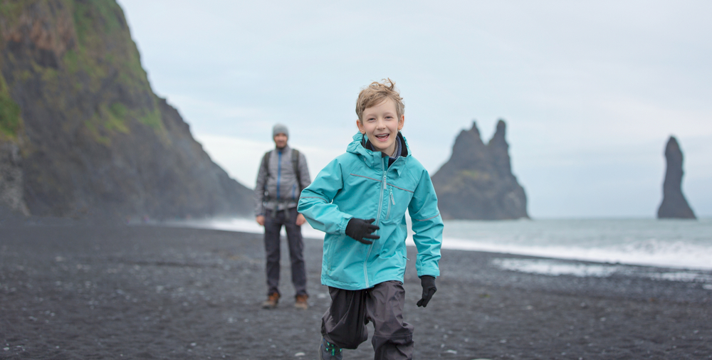 A kid in gloves, a jacket and waterproof pants runs toward the camera and away from his dad on a black sand beach.