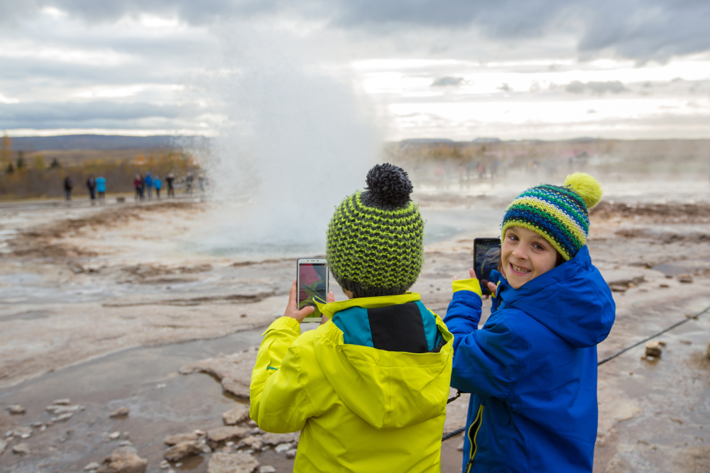 Two kids in jackets and knot beanies take photos on their phones of a hot spring in Iceland, comfortable since they knew what to wear in Iceland for kids