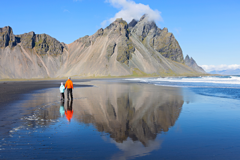 A father and son walk down a glassy black sand beach during the day, comfortable in their jackets because they knew what to wear in Iceland for kids.