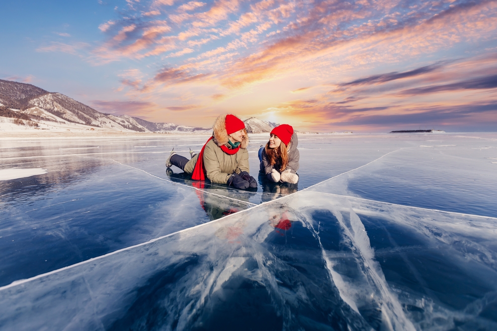 Two teens lay on the ice in Iceland in beanies, parkas and gloves during the sunrise.