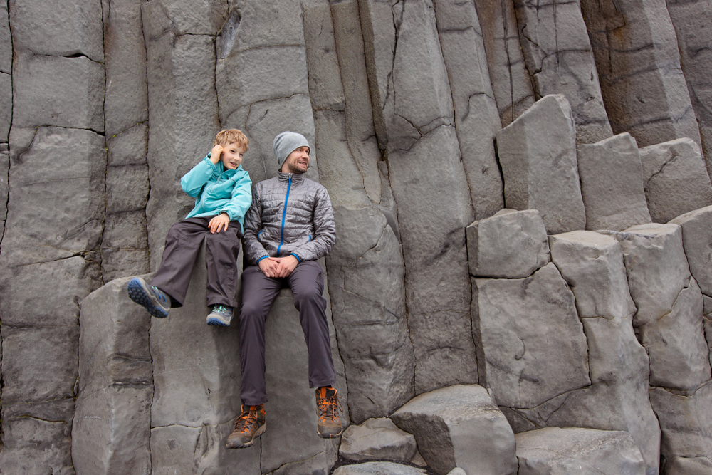 A dad and his son sit on rugged rock formations in their wind breakers and hiking boots because they knew what to wear in Iceland for kids