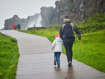 parent and child dressed in winter clothing walking along a pathway in iceland headed to an iceland waterfall in the background. the grass around them is green.