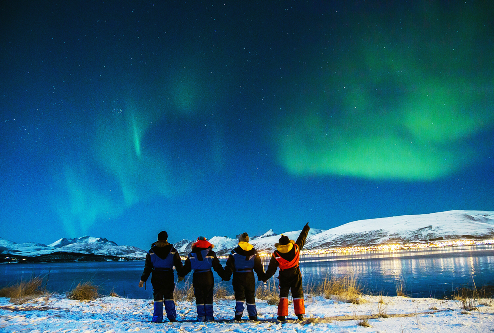 A group of tourists (a family) stand and point at the green hue in the Northern Lights. It's dark and cold, but they knew what to wear in Iceland for kids.