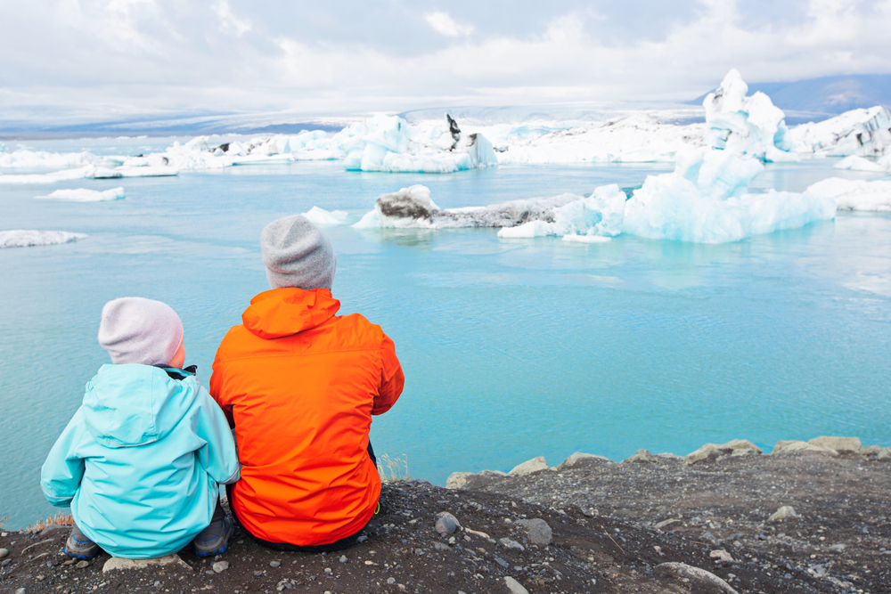 Two kids, one in an orange jacket, and one in blue, sit on the cusp of glacier water, ice in the water beyond them.