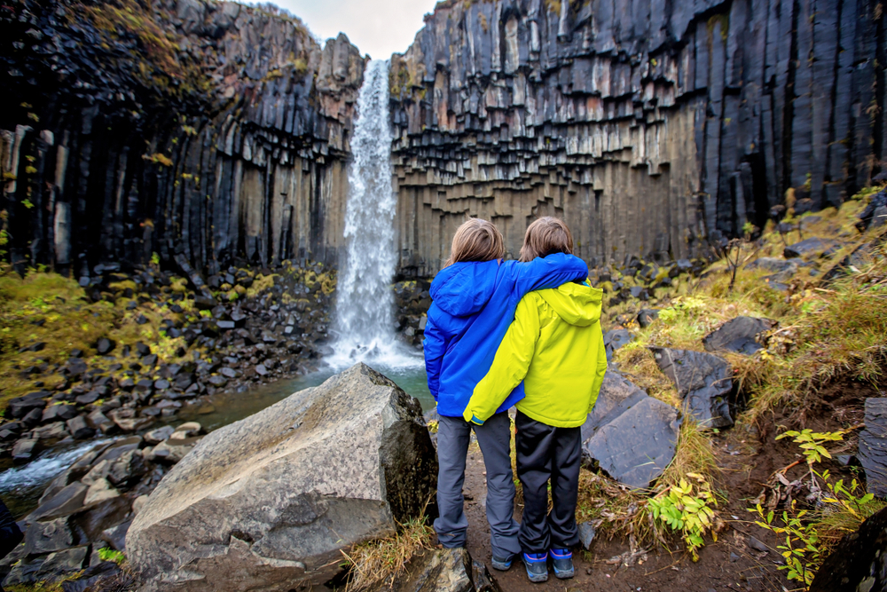 Two kids in jackets and hiking boots stand and over look a waterfall in Iceland.